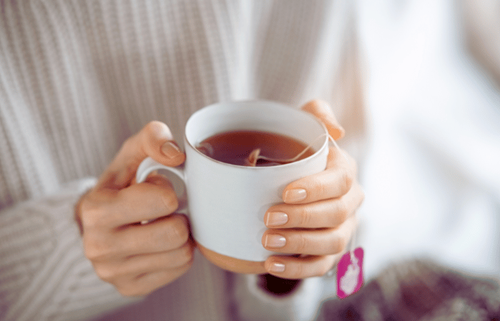 白いマグカップを両手で包み込むように持ち、温かい紅茶を楽しんでいる様子。ティーバッグのタグがカップの外に出ている。